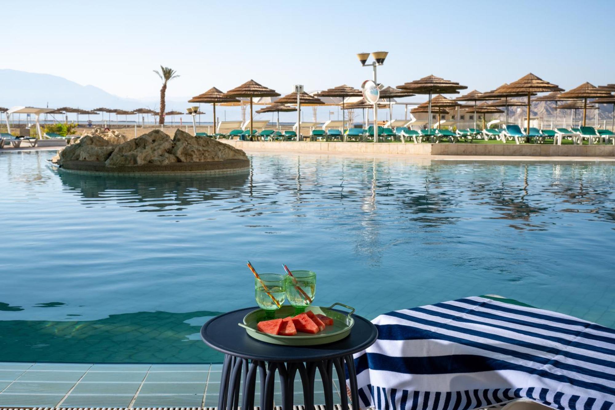 Leonardo Club Hotel Dead Sea Neve Zohar Exteriér fotografie The photo depicts a tranquil swimming pool area at a resort or hotel. In the foreground, there is a small round table with a plate containing slices of watermelon and two green drinks. The background features a calm pool with a small rock island in t