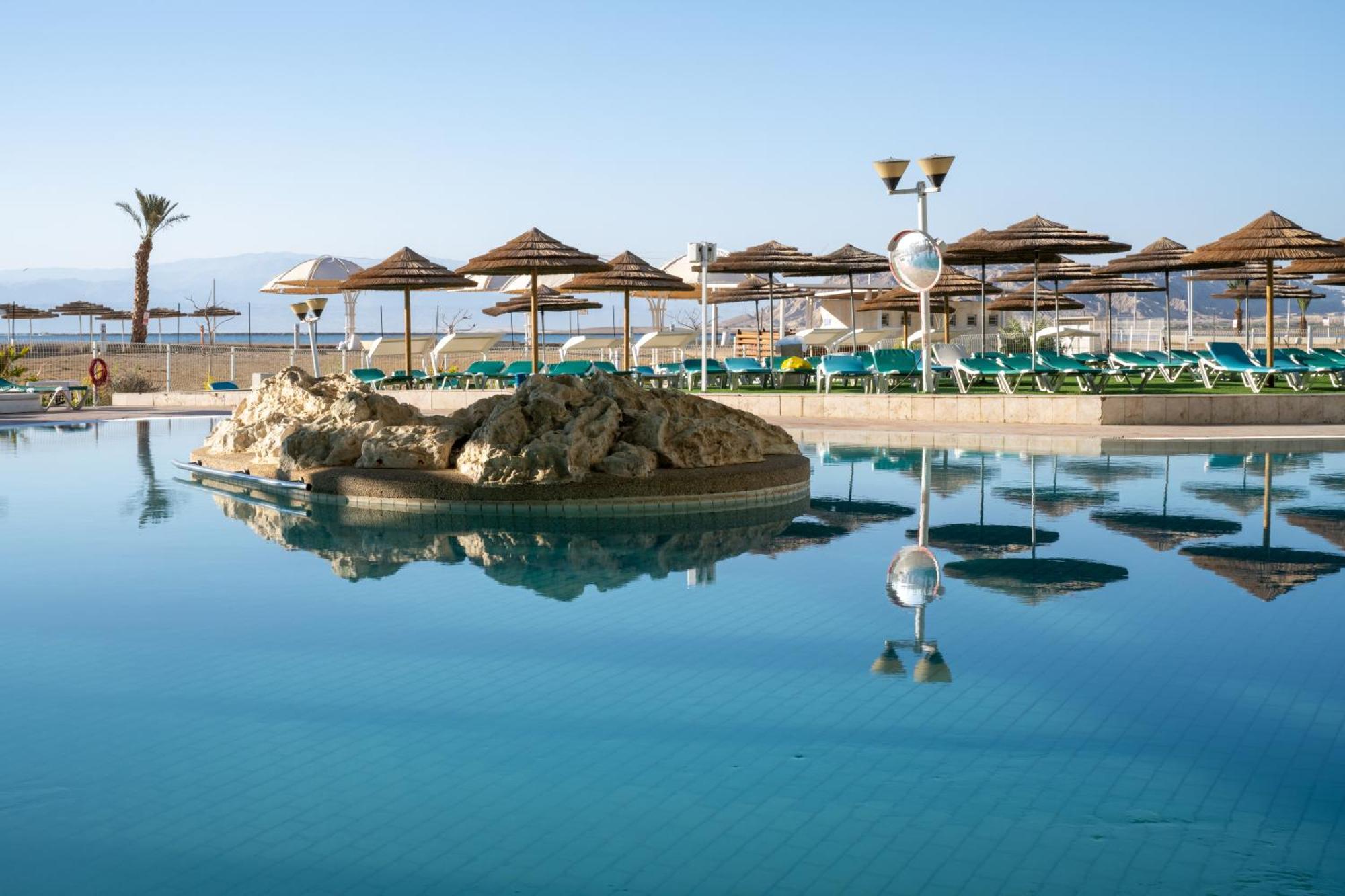 Leonardo Club Hotel Dead Sea Neve Zohar Exteriér fotografie The photo shows a tranquil swimming pool area. It features a large, reflective pool with a small rock formation in the center. Surrounding the pool are several sun loungers with green cushions and a series of thatched umbrellas providing shade. In th