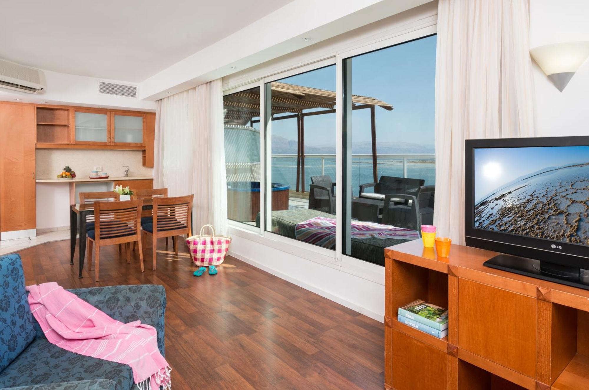 Leonardo Club Hotel Dead Sea Neve Zohar Exteriér fotografie The photo depicts a modern living space with a view. On one side, there is a comfortable seating area with a couch and a television cabinet. The TV is turned on, showing a scenic image. 

In the background, there is a dining area with a wooden table 